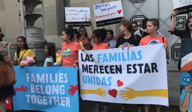 A group of adults and children standing while holding signs that read, “Families Belong Together” in English and Spanish.